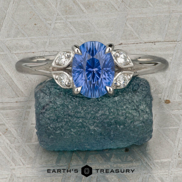 The "Hazel" ring in platinum with 1.10-carat Ceylon sapphire The "Hazel" ring in platinum with a 1.10-carat Ceylon sapphire is shown upright on a textured grey surface, resting atop a rough blue stone. “EARTH’S TREASURY” is displayed at the bottom of the image.