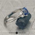 The "Hazel" ring in platinum with a 1.10-carat Ceylon sapphire is shown upright on a textured grey surface, resting atop a rough blue stone. “EARTH’S TREASURY” is displayed at the bottom of the image.