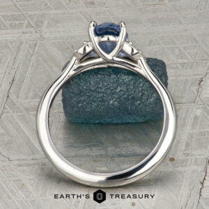 The "Hazel" ring in platinum with a 1.10-carat Ceylon sapphire is shown upright on a textured grey surface, resting atop a rough blue stone. “EARTH’S TREASURY” is displayed at the bottom of the image.