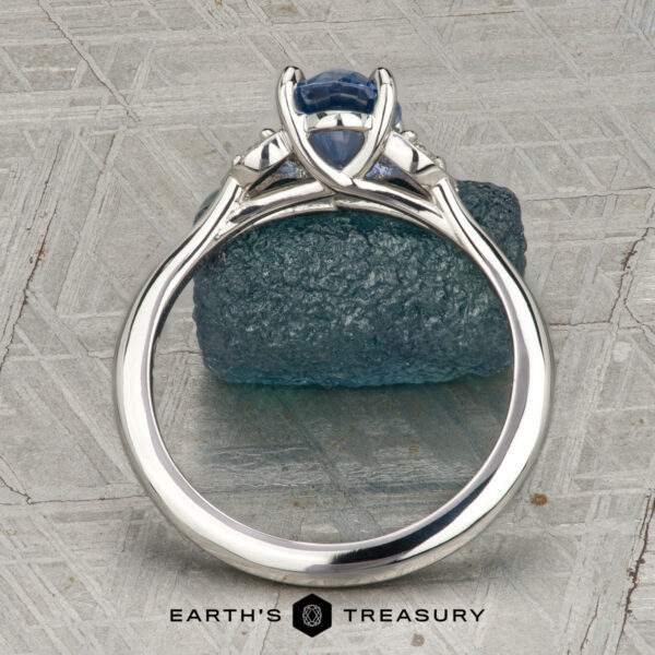 The "Hazel" ring in platinum with 1.10-carat Ceylon sapphire The "Hazel" ring in platinum with a 1.10-carat Ceylon sapphire is shown upright on a textured grey surface, resting atop a rough blue stone. “EARTH’S TREASURY” is displayed at the bottom of the image.