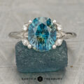 Dotson, Sean and Powers, Sarah 1: An oval blue gemstone Hazel ring with side diamonds set in a silver split-shank band. Displayed on a rough blue stone featuring the “Earth’s Treasury” logo. A unique engagement ring radiating natural elegance.