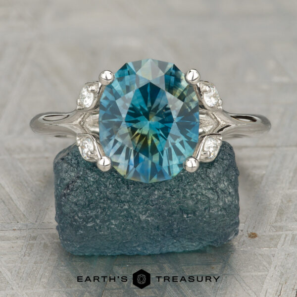 The "Hazel" ring in platinum with 3.25-carat Montana sapphire Dotson, Sean and Powers, Sarah 1: An oval blue gemstone Hazel ring with side diamonds set in a silver split-shank band. Displayed on a rough blue stone featuring the “Earth’s Treasury” logo. A unique engagement ring radiating natural elegance.