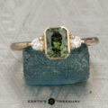 The "Evadne" ring in 14K yellow gold with a 0.95-carat Australian sapphire and two round white stones is displayed on a textured blue surface, with the text "EARTH'S TREASURY" at the bottom.
