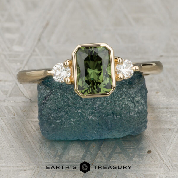 The "Evadne" ring in 14K yellow gold with 0.95-carat Australian sapphire The "Evadne" ring in 14K yellow gold with a 0.95-carat Australian sapphire and two round white stones is displayed on a textured blue surface, with the text "EARTH'S TREASURY" at the bottom.
