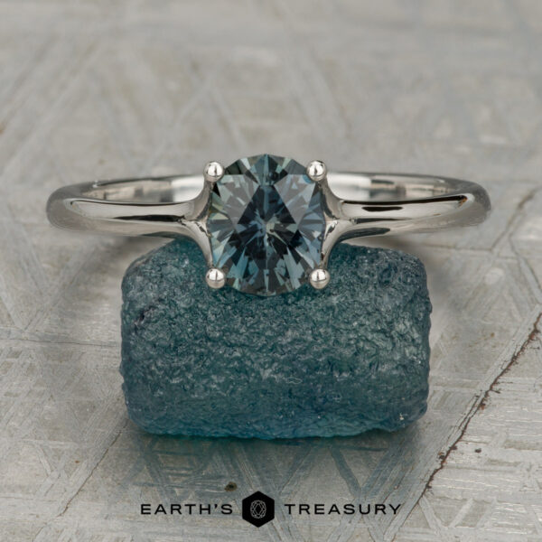 The "Phoebe" ring in platinum with 1.12-carat Songea sapphire The "Phoebe" ring in platinum, featuring a 1.12-carat Songea sapphire, sits atop a rough blue crystal on a textured surface. "Earth's Treasury" is written below.