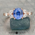 The Cunningham, Brian ring features a Cattleya-inspired rose gold band with an oval blue center stone, accented by four marquise and two round clear stones, set on dark mineral rock for a striking jewelry statement.