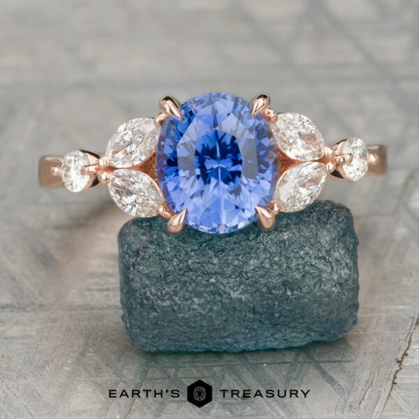 The "Cattleya" ring in 14K rose gold with 2.95-carat Ceylon sapphire The Cunningham, Brian ring features a Cattleya-inspired rose gold band with an oval blue center stone, accented by four marquise and two round clear stones, set on dark mineral rock for a striking jewelry statement.