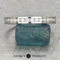 The Classic Channel-Set Band in platinum, featuring a row of round diamonds, stands upright on a rough blue gemstone against a geometric background with the Earth’s Treasury logo below.