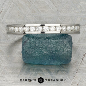 The Classic Channel-Set Band in platinum, featuring a row of round diamonds, stands upright on a rough blue gemstone against a geometric background with the Earth’s Treasury logo below.