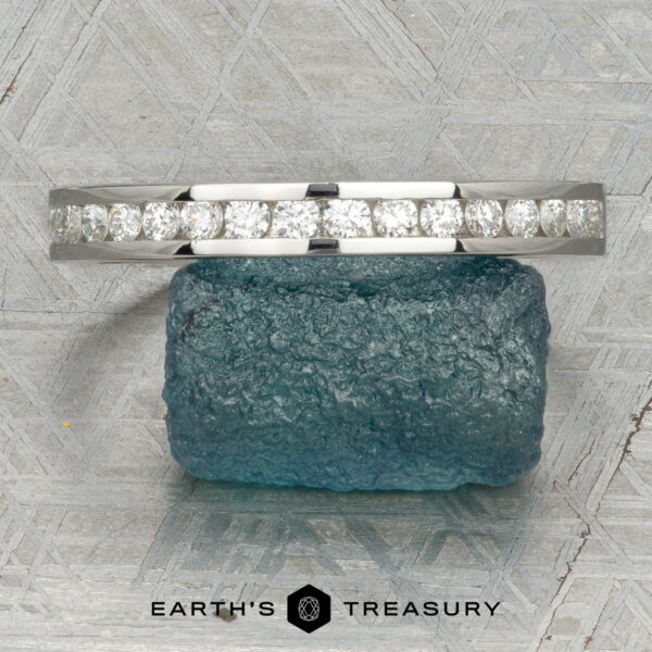 The Classic Channel-Set Band in platinum The Classic Channel-Set Band in platinum, featuring a row of round diamonds, stands upright on a rough blue gemstone against a geometric background with the Earth’s Treasury logo below.