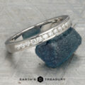 The Classic Channel-Set Band in platinum, featuring a row of round diamonds, stands upright on a rough blue gemstone against a geometric background with the Earth’s Treasury logo below.