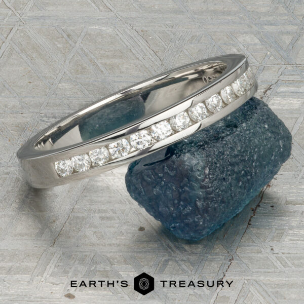 The Classic Channel-Set Band in platinum The Classic Channel-Set Band in platinum, featuring a row of round diamonds, stands upright on a rough blue gemstone against a geometric background with the Earth’s Treasury logo below.