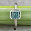 The Johnson, Dalton and Murphy, Erin solitaire silver ring showcases an emerald-cut blue gemstone, elegantly displayed on a light green surface with the Earth's Treasury logo at the bottom.