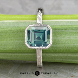 The Johnson, Dalton and Murphy, Erin solitaire silver ring showcases an emerald-cut blue gemstone, elegantly displayed on a light green surface with the Earth's Treasury logo at the bottom.