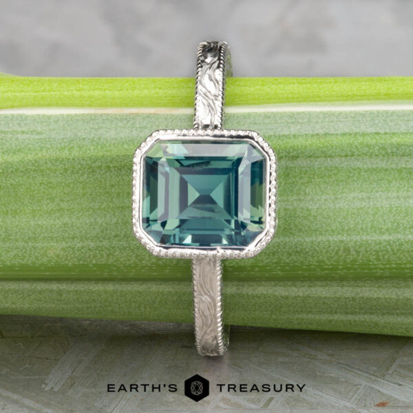 The "Niobe" ring in platinum with 2.35-carat Ceylon sapphire The Johnson, Dalton and Murphy, Erin solitaire silver ring showcases an emerald-cut blue gemstone, elegantly displayed on a light green surface with the Earth's Treasury logo at the bottom.