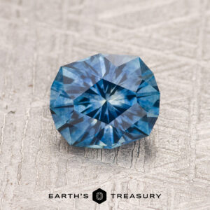 A 1.37-carat Teal Blue Montana Sapphire (Heated), faceted and round, is shown on a textured light surface with "Earth's Treasury" written beneath the stone.