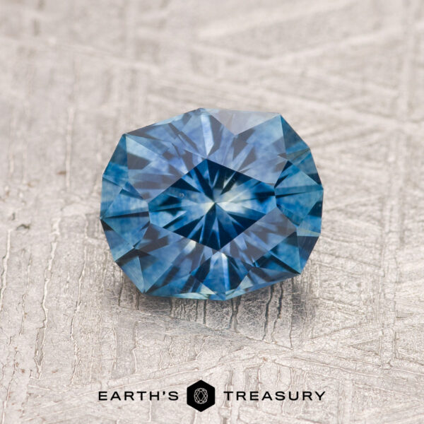 1.37-Carat Teal Blue Montana Sapphire (Heated) A 1.37-carat Teal Blue Montana Sapphire (Heated), faceted and round, is shown on a textured light surface with "Earth's Treasury" written beneath the stone.