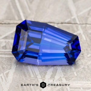 A breathtaking 3.89-carat Medium Blue Tanzanite (Heated) gemstone with an irregular step-cut shape rests on a textured gray surface, with the text "Earth's Treasury" beneath it.