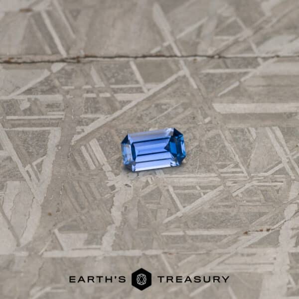 A stunning 0.38-Carat Cornflower Blue Yogo Sapphire in a rectangular cut placed on a textured, metallic surface. The logo "Earth's Treasury" is displayed at the bottom center of the image.