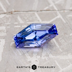 A 1.23-Carat Light Violet-Blue Tanzanite (Heated) with a faceted hexagonal shape rests on a textured silver surface. The gemstone’s striking light violet-blue hues catch the light beautifully, while the "Earth's Treasury" logo appears at the bottom of the image.