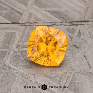 A 0.77-Carat Spessartine Garnet gemstone, featuring a square cut with intricate facets, rests on a textured gray surface.