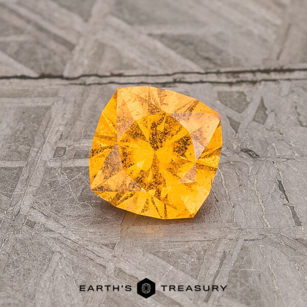 0.77-Carat Spessartine Garnet A 0.77-Carat Spessartine Garnet gemstone, featuring a square cut with intricate facets, rests on a textured gray surface.