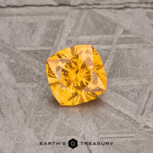 A vibrant 0.56-carat Spessartine Garnet gleams on a textured gray surface, with "Earth's Treasury" elegantly inscribed below.