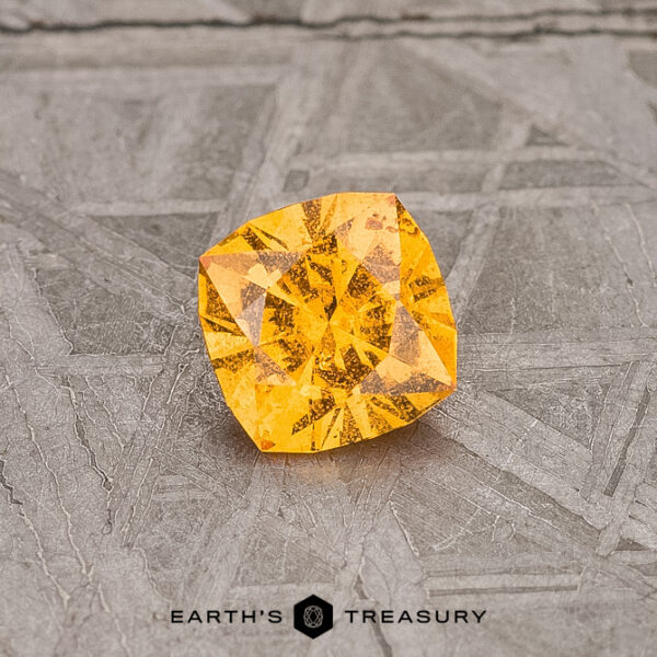 0.56-Carat Spessartine Garnet A vibrant 0.56-carat Spessartine Garnet gleams on a textured gray surface, with "Earth's Treasury" elegantly inscribed below.