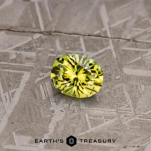 A 2.74-carat lime green Australian sapphire, expertly cut in a round, brilliant shape, is placed on a patterned surface. Its facets reflect light beautifully, creating a captivating sparkling effect. Below the gemstone, the text "Earth's Treasury" can be seen along with a logo that features a geometric design.