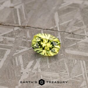 A 2.74-carat lime green Australian sapphire, brilliantly cut into an oval shape, is displayed on a textured gray surface. The gemstone exhibits a clear, sparkly appearance with facets that reflect light. The text "EARTH'S TREASURY" is visible at the bottom center of the image.