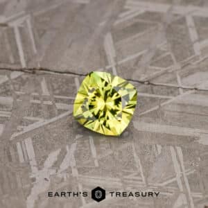 A faceted, square-shaped 2.74-carat lime green Australian sapphire rests on a textured, metallic surface. The surface has an intricate, geometric pattern with various intersecting lines and shapes. The text "EARTH'S TREASURY" appears at the bottom of the image.
