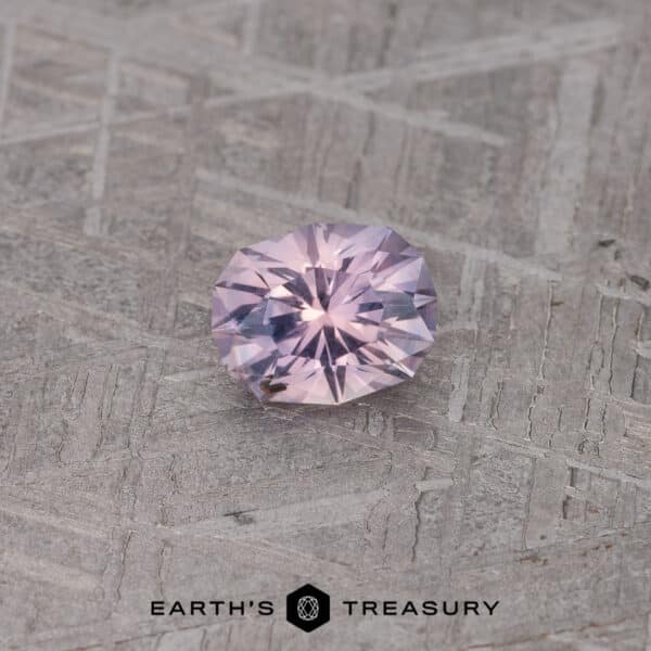 0.83-Carat Lavender-Pink Montana Saphire A sparkling, round-cut 0.83-carat lavender-pink Montana sapphire is displayed on a geometric-patterned grey surface. The gemstone's facets reflect light, showcasing its clarity and intricate cuts. The logo and text "Earth's Treasury" are visible at the bottom of the image.
