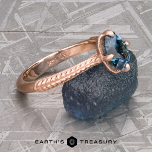 The 2022-01-10 11-10-57 (B,Radius1,Smoothing1)-1200-BW ring features a rose gold Demeter Solitaire design with a blue gemstone and engraved band, set on a rough blue stone atop a textured gray surface with the Earth's Treasury logo.