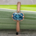 The 2022-01-10 11-17-33 (B,Radius1,Smoothing1)-1200-BW ring, featuring a blue gemstone in rose gold, is displayed vertically on a green plant stem. Below the ring, the Earth's Treasury logo is visible.
