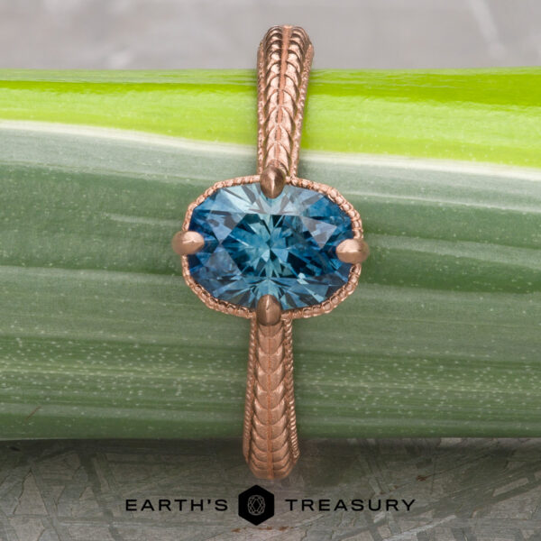 The “Demeter” Ring in 14k rose gold with 1.18-Carat Montana Sapphire The 2022-01-10 11-17-33 (B,Radius1,Smoothing1)-1200-BW ring, featuring a blue gemstone in rose gold, is displayed vertically on a green plant stem. Below the ring, the Earth's Treasury logo is visible.
