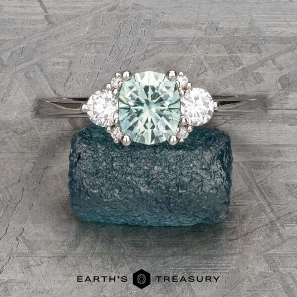 The "Aurelia" ring in 14k white gold with 1.05-carat Montana sapphire The "Aurelia" ring in 14k white gold showcases a central light green Montana sapphire weighing 1.05 carats, accented by two clear side stones, all set against an elegantly textured finish.