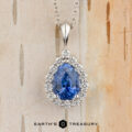 The Cheng, Yuhe pendant features a pear-shaped blue gemstone framed by a diamond halo on a silver chain, elegantly displayed against a light wood background.