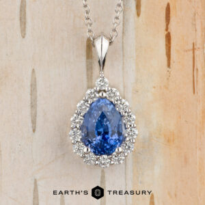 The Cheng, Yuhe pendant features a pear-shaped blue gemstone framed by a diamond halo on a silver chain, elegantly displayed against a light wood background.