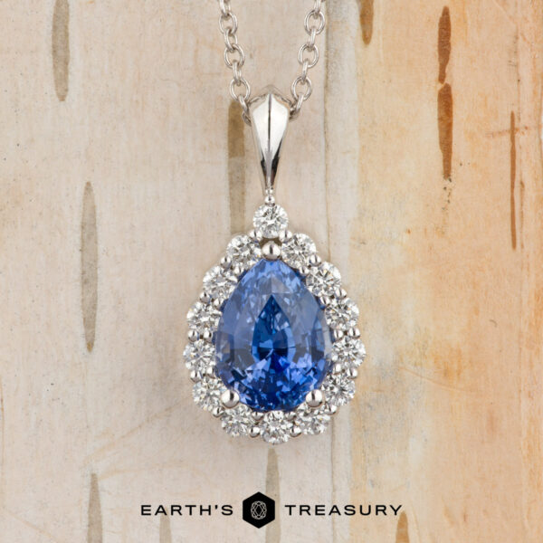 The BYO Classic Diamond Halo Pendant in 18K white gold with 1.64-carat Ceylon sapphire The Cheng, Yuhe pendant features a pear-shaped blue gemstone framed by a diamond halo on a silver chain, elegantly displayed against a light wood background.