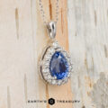 The BYO Classic Diamond Halo Pendant in 18K white gold features a 1.64-carat pear-shaped Ceylon sapphire and round diamonds on a silver chain, set against a light wooden background.