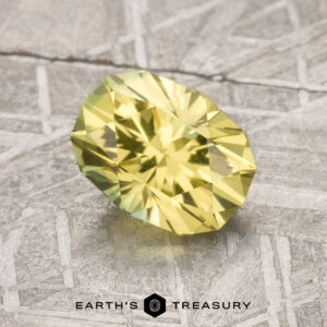 A 1.72-carat Lemon Yellow Australian Sapphire, faceted in an oval shape, is shown on a textured gray surface with the "Earth's Treasury" logo beneath it.