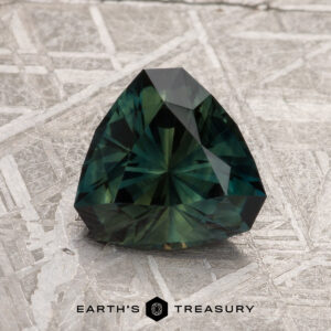 A faceted 2.16-carat dark green Australian sapphire with a trillion cut rests on a textured metallic surface. The text "Earth's Treasury" appears at the bottom.