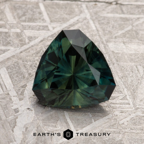 2.16-Carat Dark Green Australian Sapphire A faceted 2.16-carat dark green Australian sapphire with a trillion cut rests on a textured metallic surface. The text "Earth's Treasury" appears at the bottom.
