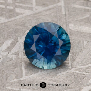 A 1.35-carat Rich Blue Montana Sapphire (Heated), round and faceted, is shown on a textured metallic surface with "EARTH'S TREASURY" featured at the bottom.