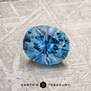 A stunning 1.22-carat medium blue Montana sapphire, expertly cut into an oval shape, rests on a textured surface resembling finely etched metal. The gemstone is elegantly highlighted with the "Earth's Treasury" logo and text centered at the bottom of the image.