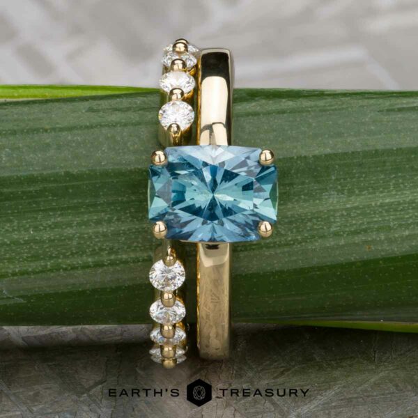 The "Freya" ring in 14k yellow gold with 2.27-Carat Montana Sapphire, alongside the "Andelle" band in 14k yellow gold The “Freya” ring in 14k yellow gold with a 2.27-carat Montana sapphire is paired with the “Andelle” open band in 14k yellow gold, both displayed on a green leaf.