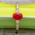 The "Galina" ring in 18k yellow gold with a 1.34-carat Spinel has a round red center stone and marquise clear accents, displayed on a green surface above the "Earth's Treasury" logo.