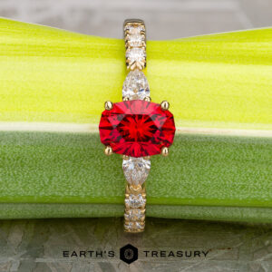The "Galina" ring in 18k yellow gold with a 1.34-carat Spinel has a round red center stone and marquise clear accents, displayed on a green surface above the "Earth's Treasury" logo.