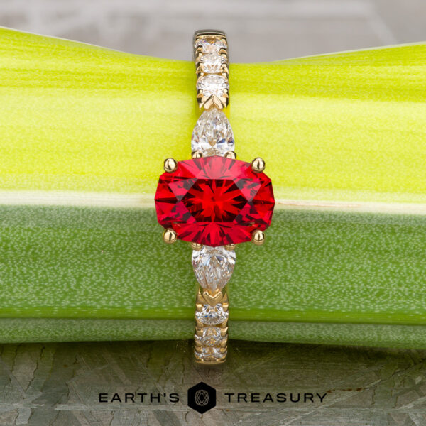 The "Galina" ring in 18k yellow gold with 1.34-carat Spinel The "Galina" ring in 18k yellow gold with a 1.34-carat Spinel has a round red center stone and marquise clear accents, displayed on a green surface above the "Earth's Treasury" logo.
