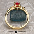 The "Galina" ring in 18k yellow gold with a 1.34-carat Spinel stands upright before a rough blue stone on a textured gray surface, with the Earth's Treasury logo at the bottom.